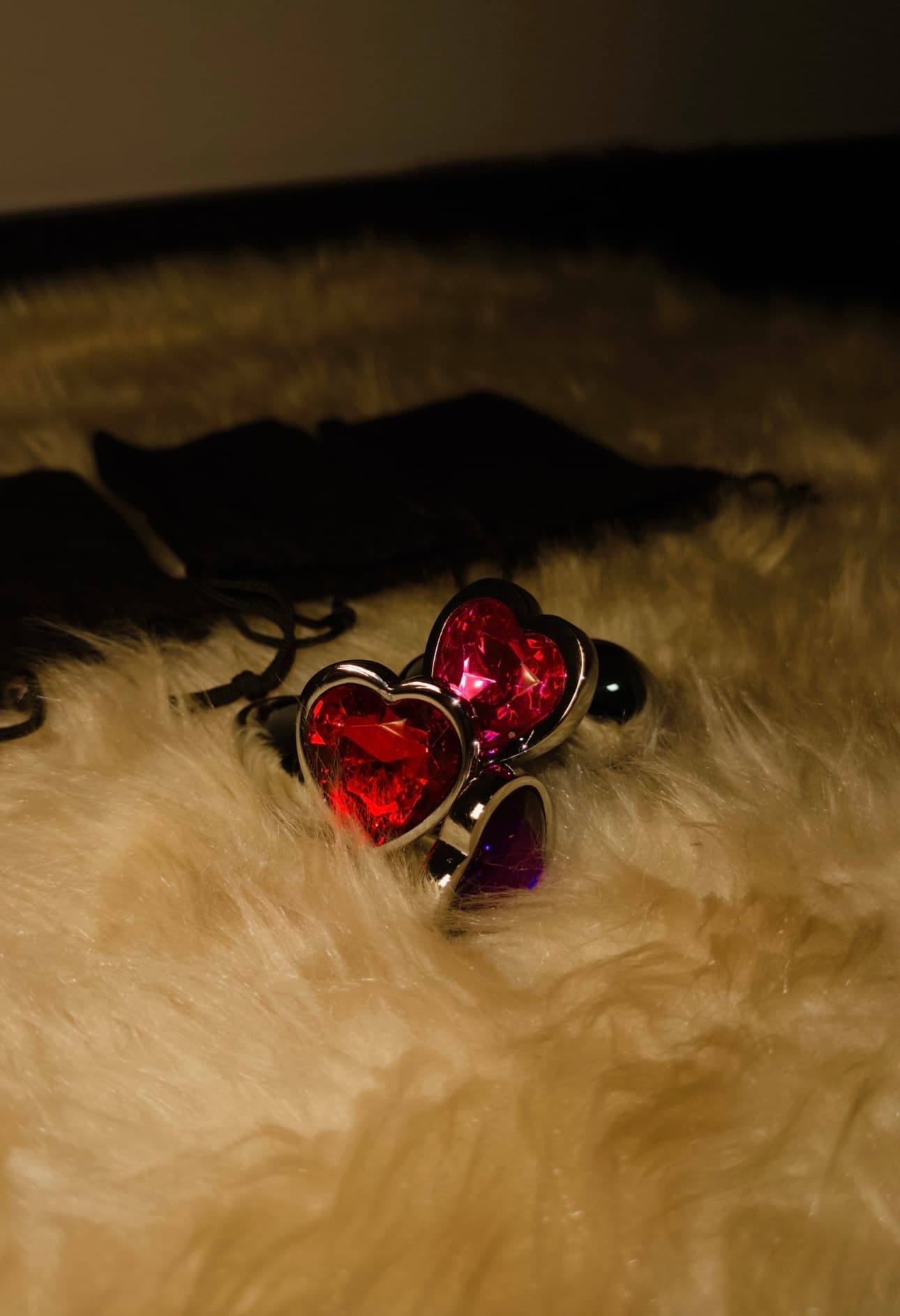 Plugs de metal com pedras em formato de coração vermelha, rosa e roxa, sobre pelúcia creme